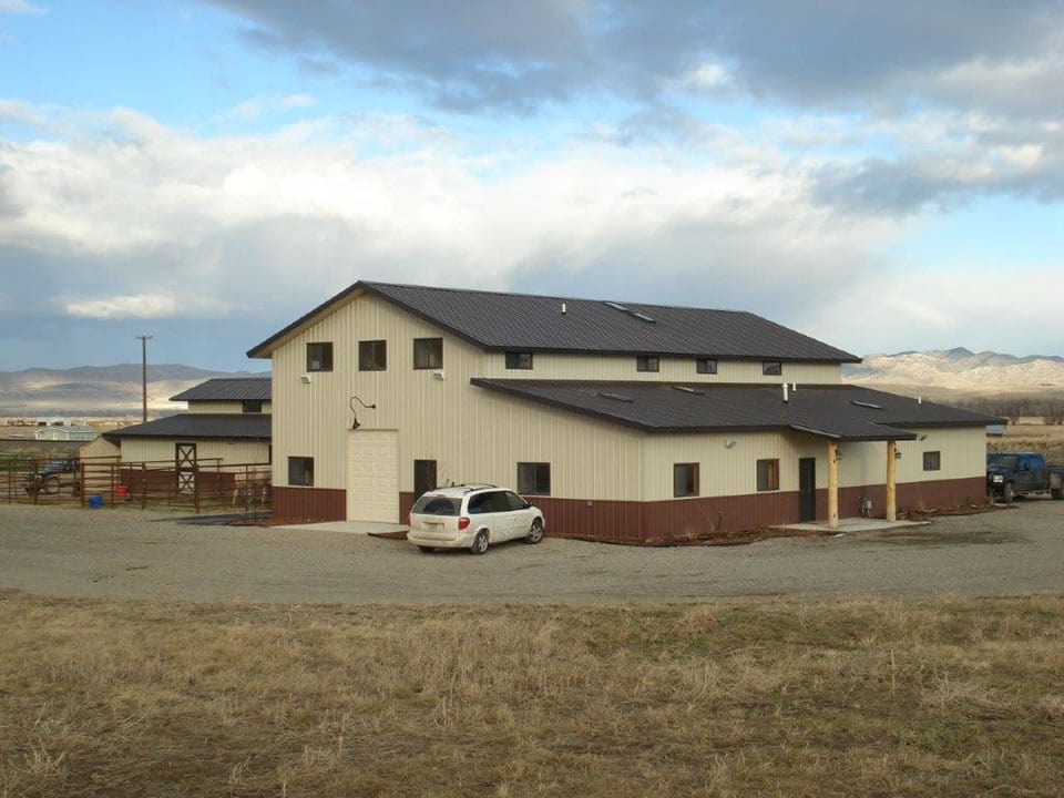Light-colored barn-like building with brown trim, small windows, and a few vehicles parked outside.