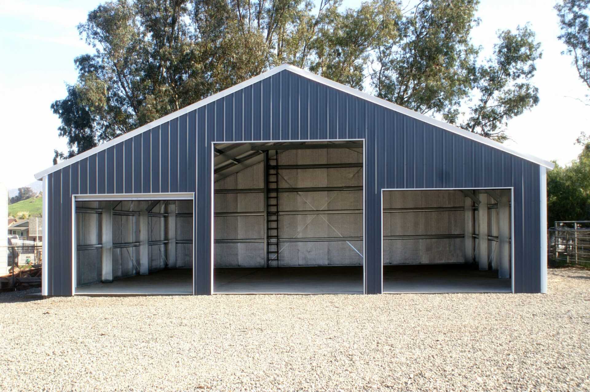 Blue metal building with three bays, gravel driveway, and a ladder inside.