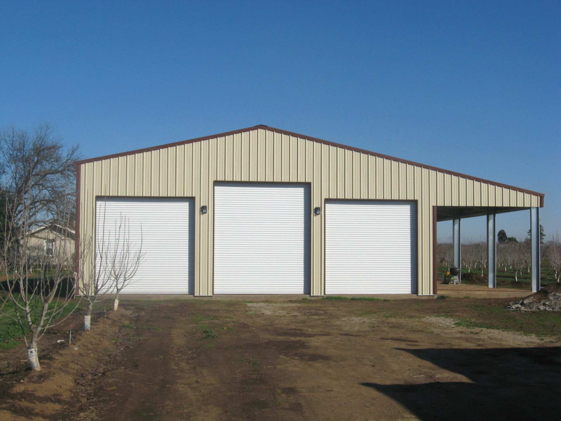 Tan metal garage with three white garage doors and a carport. Sunny day.