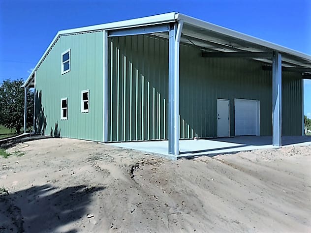 Green metal building with a covered area and open entrance, set on a dirt lot under a blue sky.