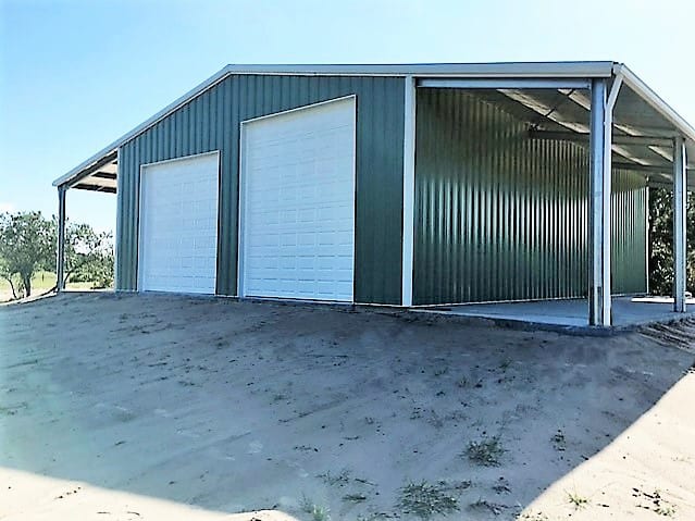 Green metal building with two garage doors and a carport in a field.
