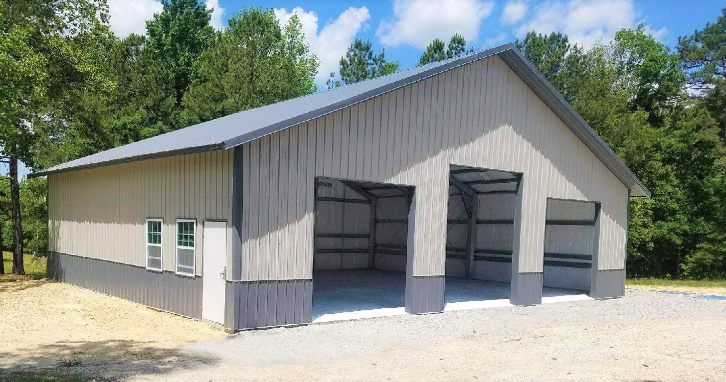 Gray metal barn with two garage bays open, on a gravel lot, with a forest background.