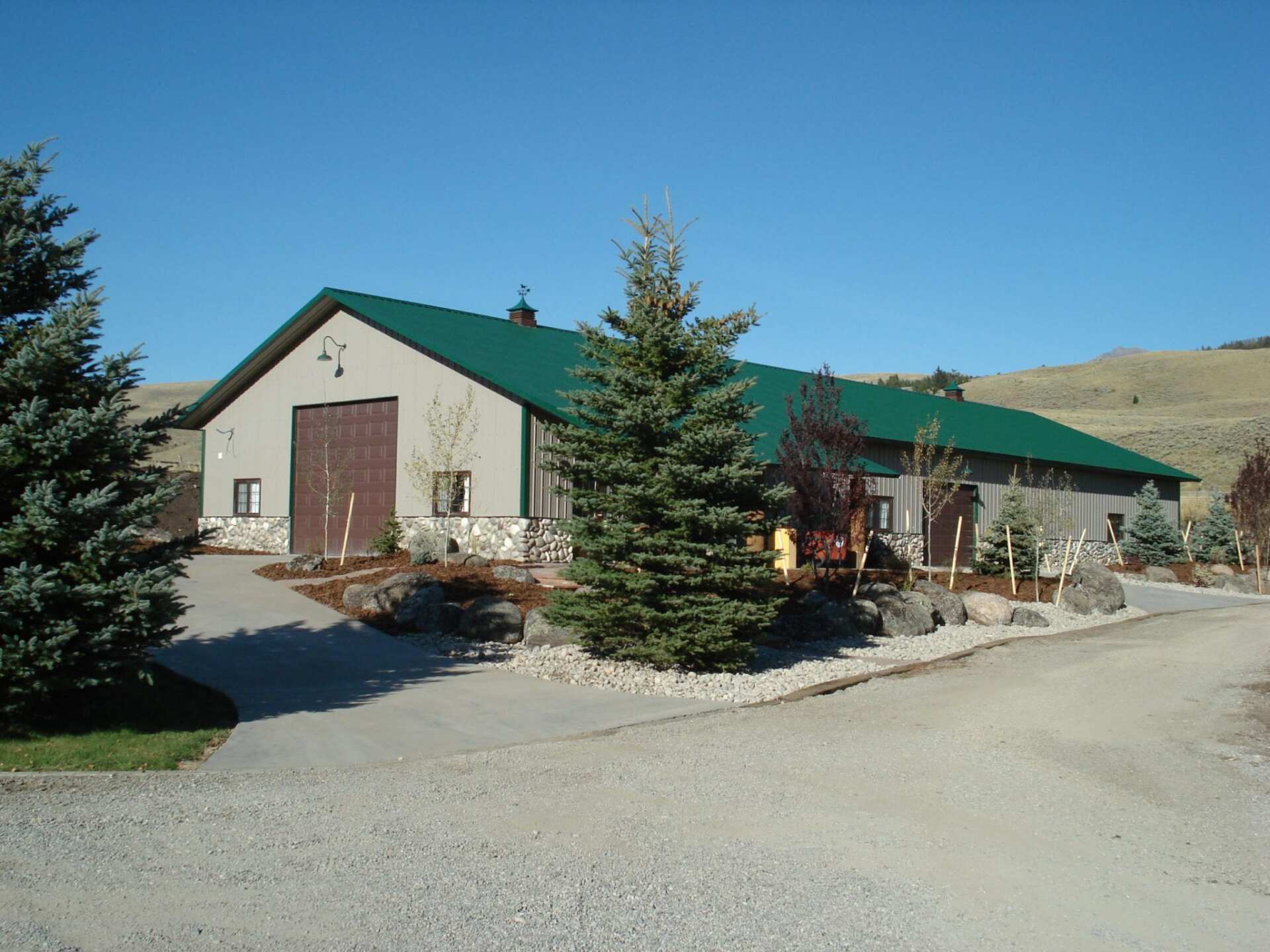 Tan building with a green roof and brown door, set in a gravel driveway with trees and a clear sky.