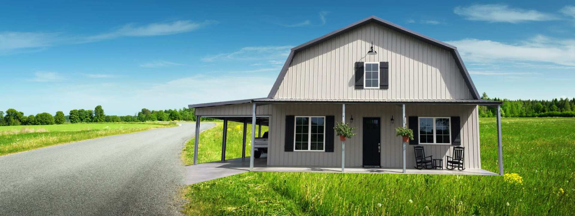A gray barn-style house with a porch, located on a green field with a road leading to it under a blue sky.