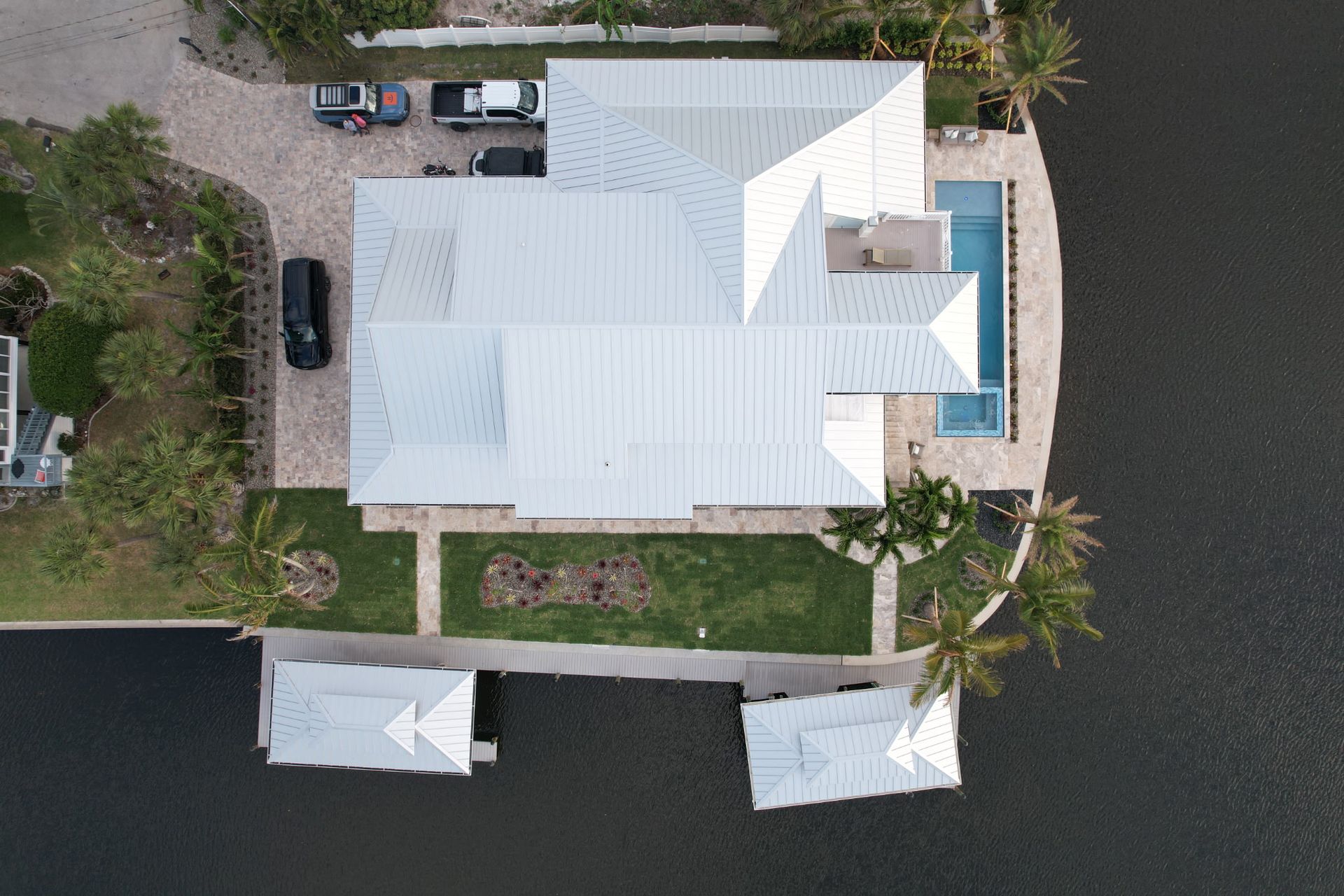 Aerial view of a white roof house with a pool and boat docks on a waterway. Cars parked in the driveway.