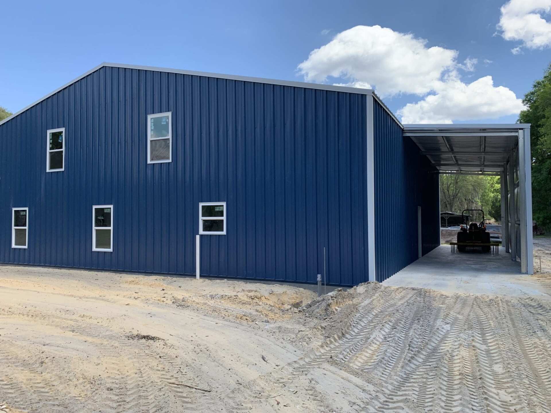 Blue metal building with carport, windows, and a sandy driveway under a cloudy sky.