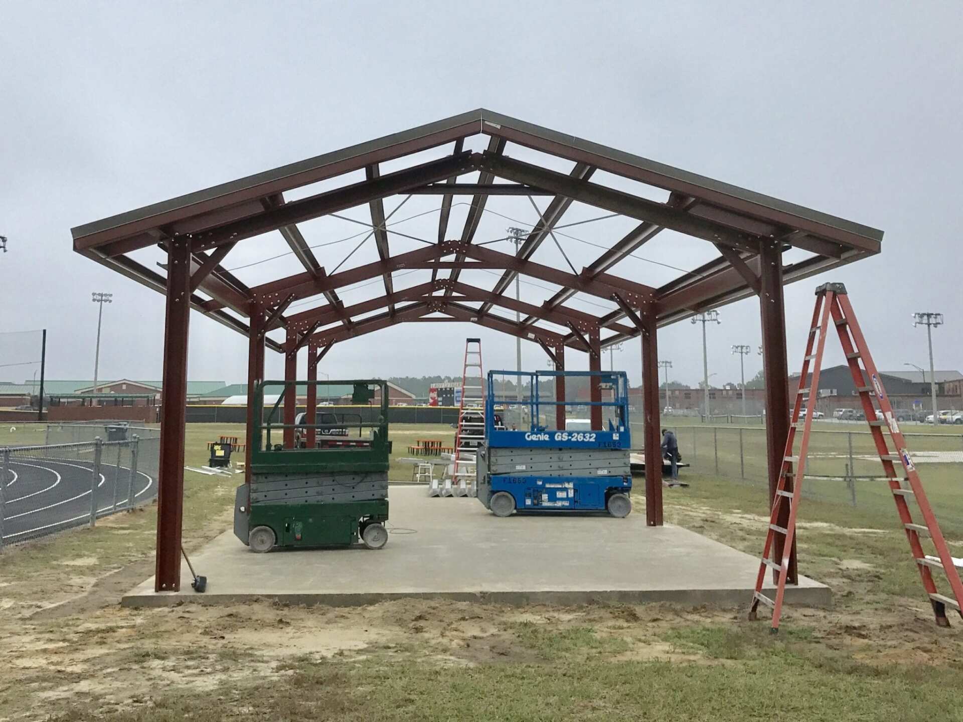 A metal pavilion under construction with two lifts, a ladder, and a sports field in the background.