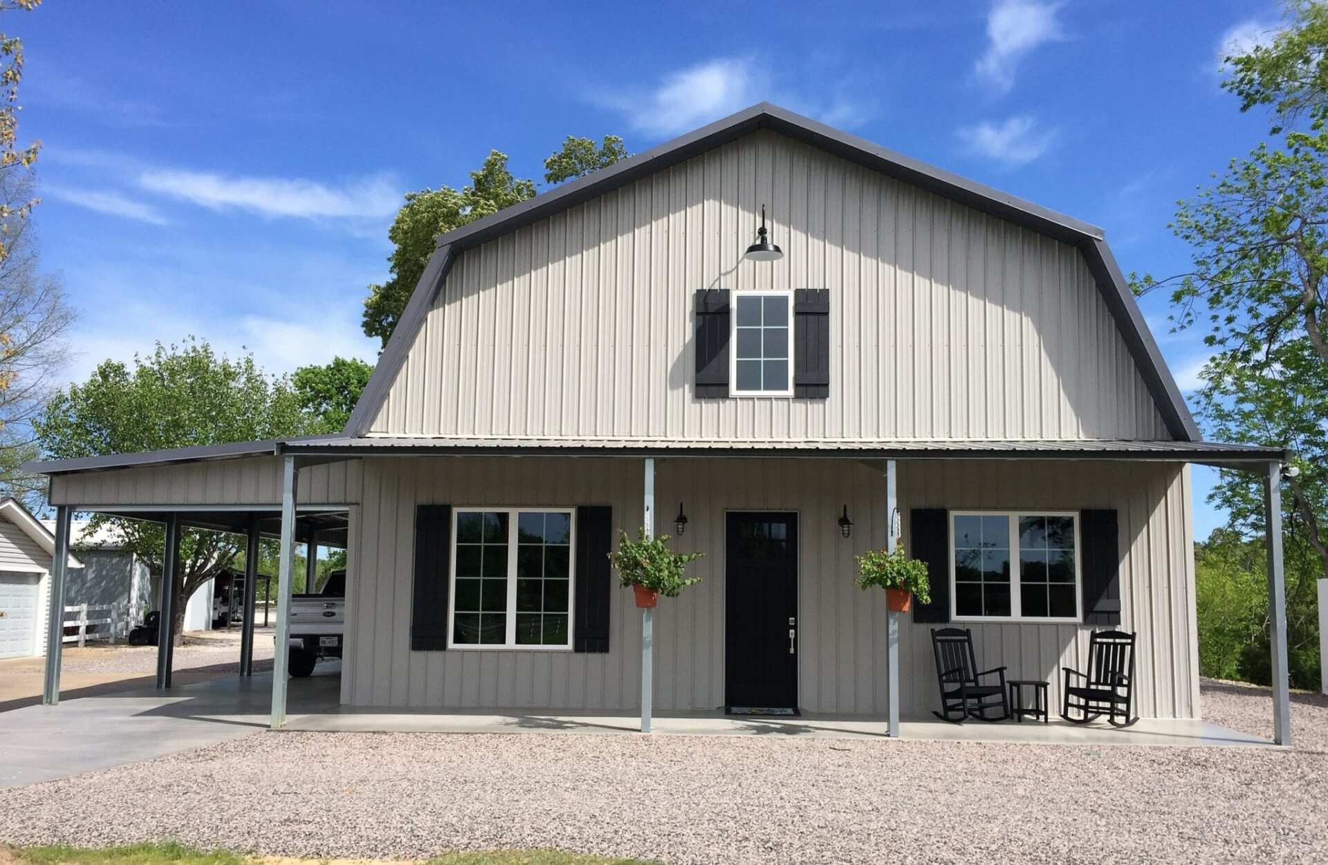 Barn-style house with porch, light gray siding, black shutters and door, gravel yard, and blue sky.