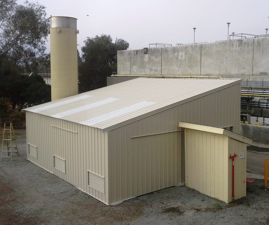 Tan industrial building with a sloped roof, connected to a smaller shed, with a smokestack in the background.