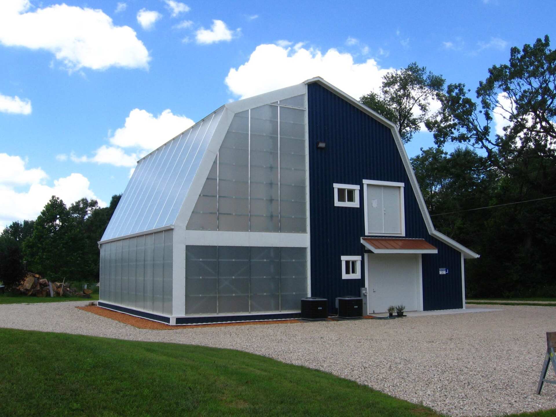 Blue barn with a large solar panel facade against a blue sky, on a gravel driveway.
