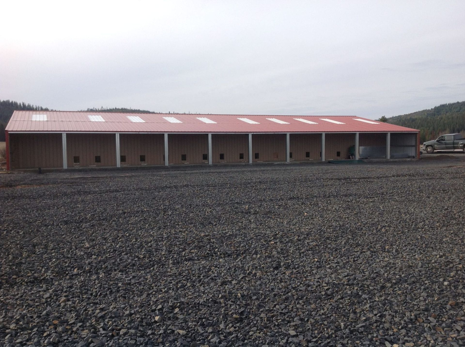 A long, tan metal building with a red roof sits on a gravel lot.