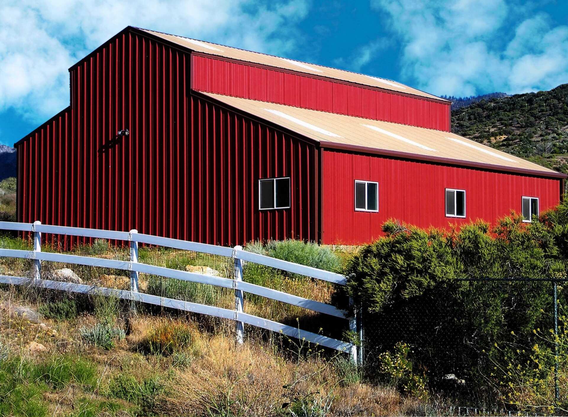 Red metal barn with a beige roof, white fence, and green bushes.