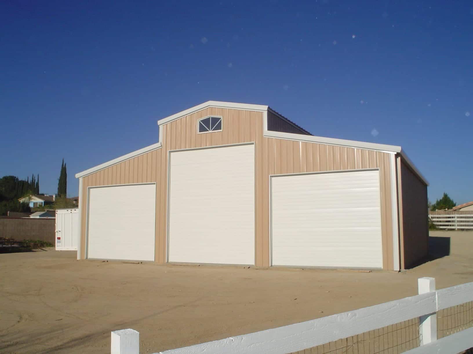 Tan and white metal barn with three garage doors under a clear blue sky.