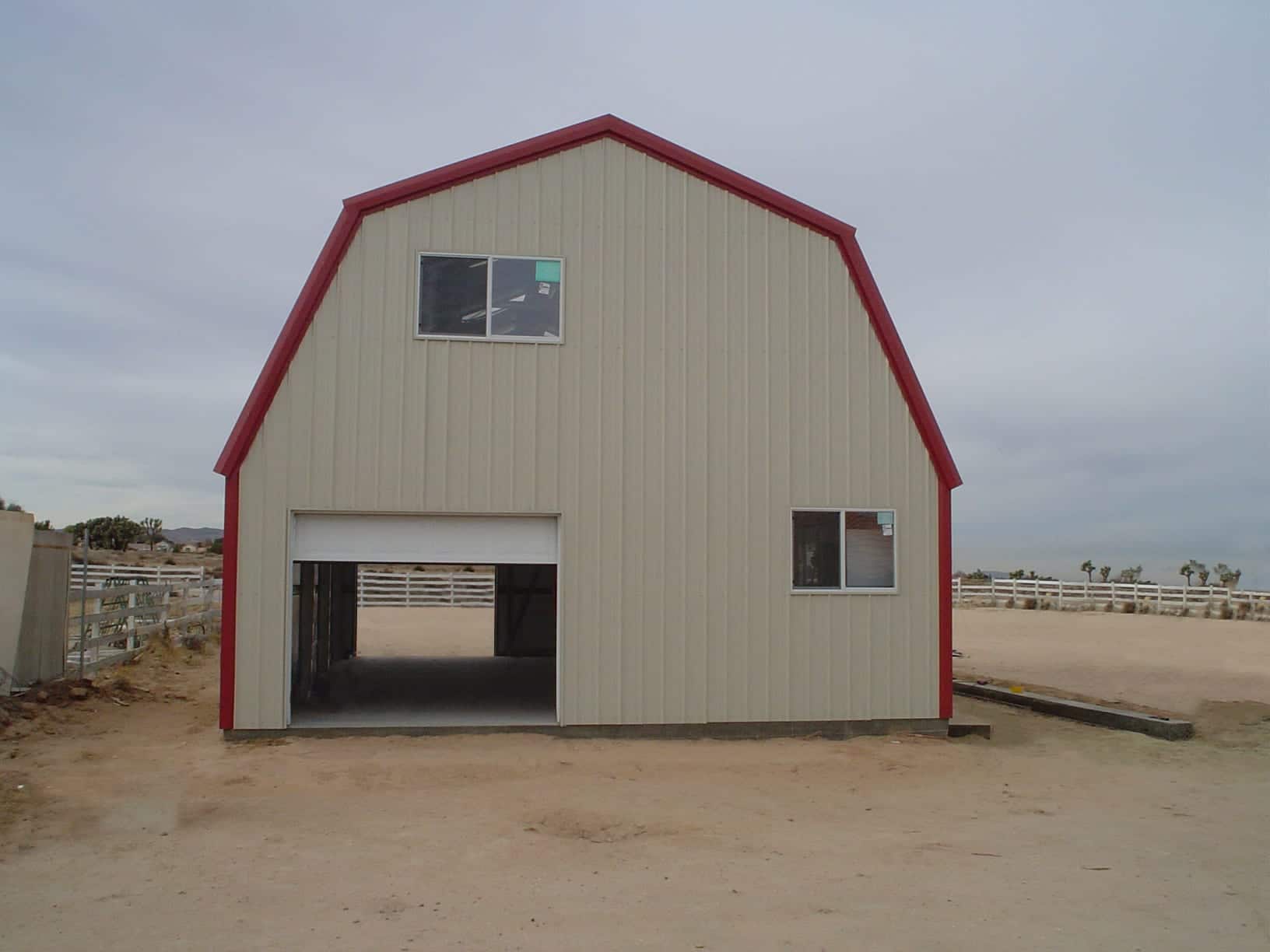 Beige barn-style metal building with red trim, open garage door, and windows under a cloudy sky.