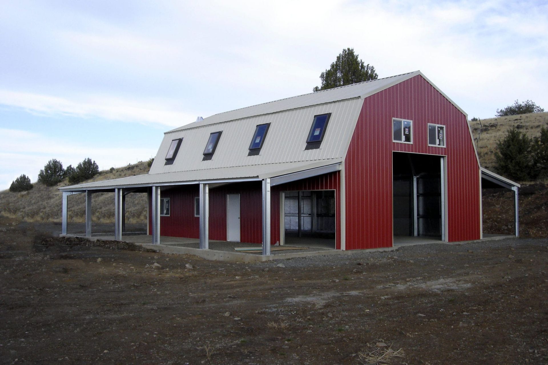 Red barn with a white roof and awnings, windows, and open doors, set on a gravel lot near a hill.
