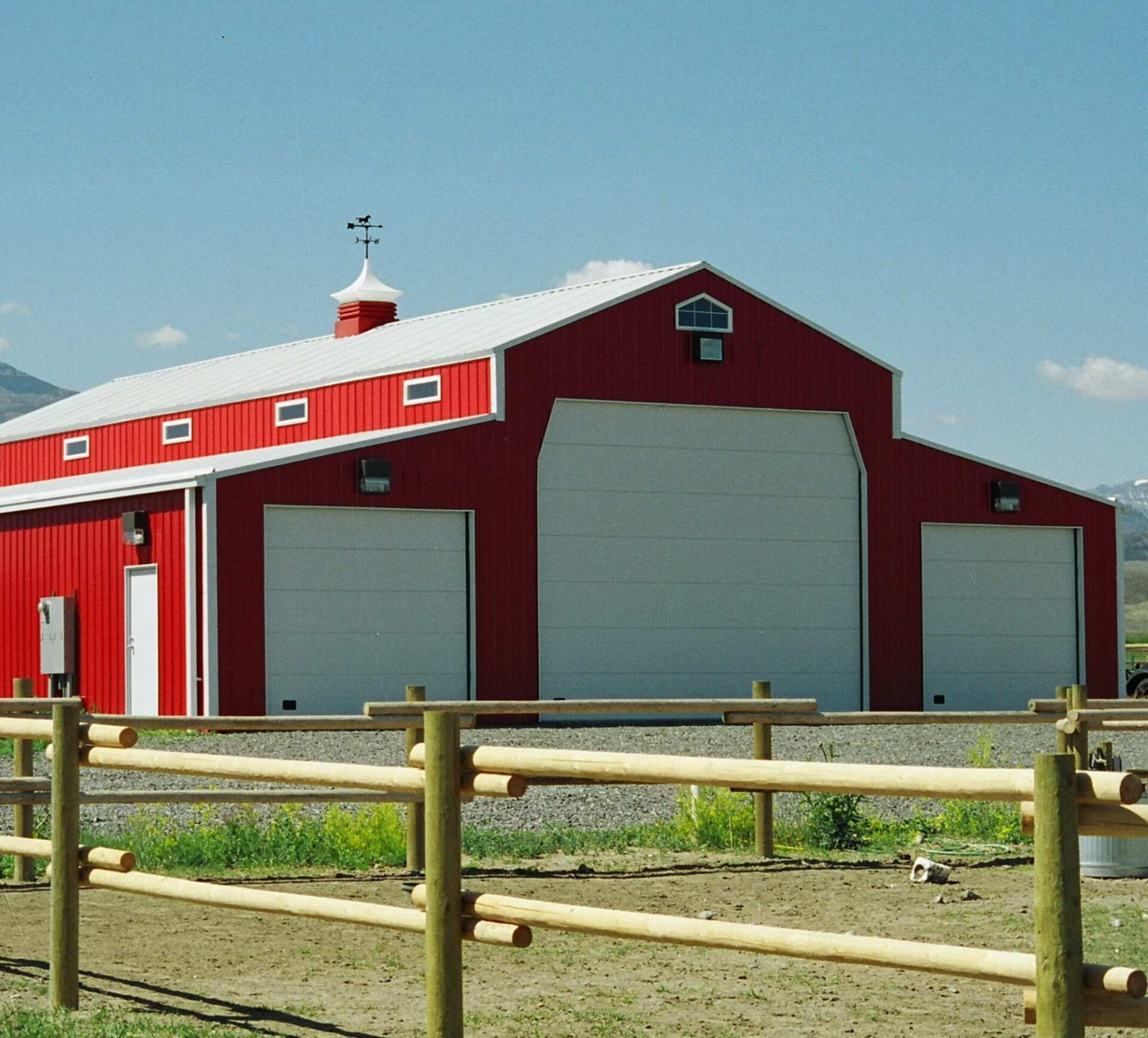 Red barn with white doors and roof, wooden fence in front, mountains in the background.