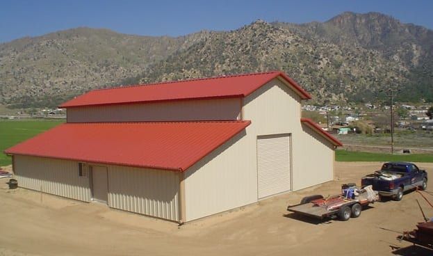 Red-roofed barn with open door, light tan exterior, mountains in background, truck and trailer parked in front.