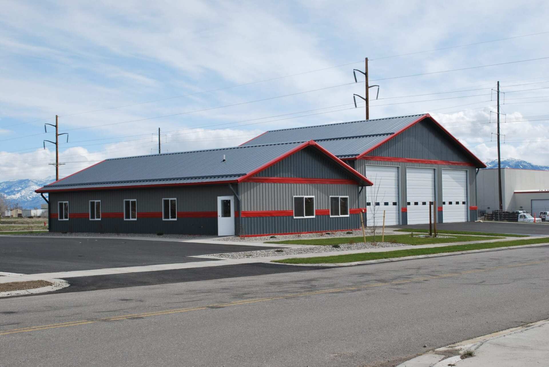 Gray metal building with red trim, featuring two garage doors and windows, under a cloudy sky.