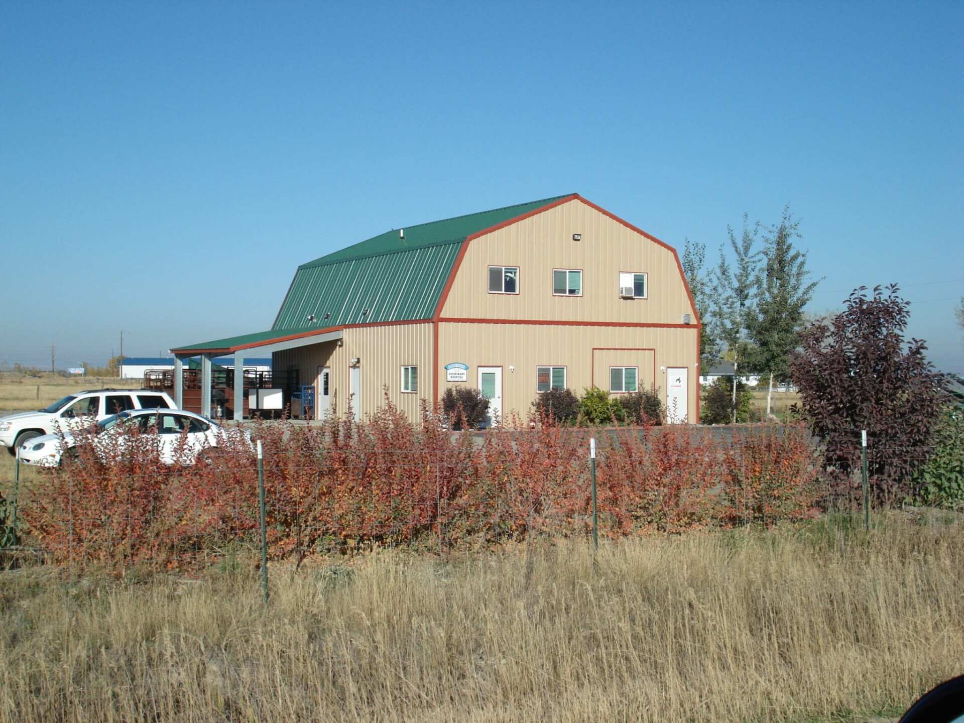 Tan barn-style building with green roof, red trim, bushes, and vehicles on a sunny day.