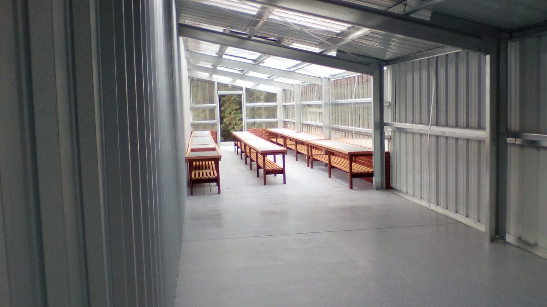 Inside a metal-walled greenhouse with long wooden tables under a translucent roof.