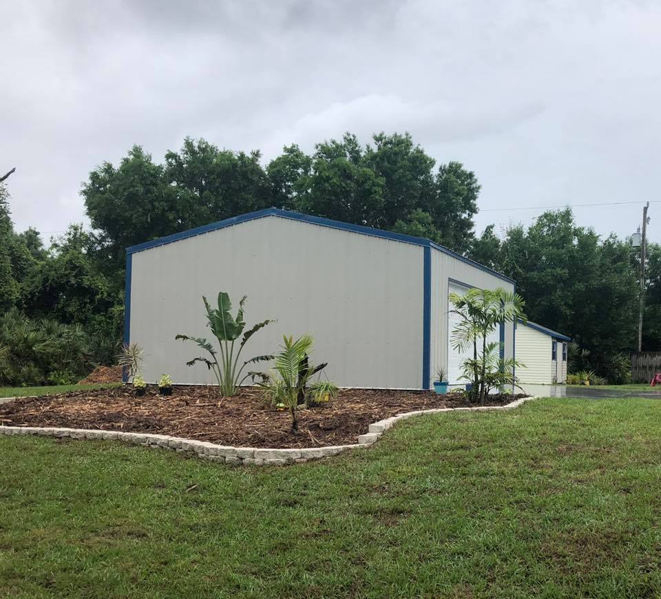 Large light-colored building with blue trim, garden bed in front, and trees behind.