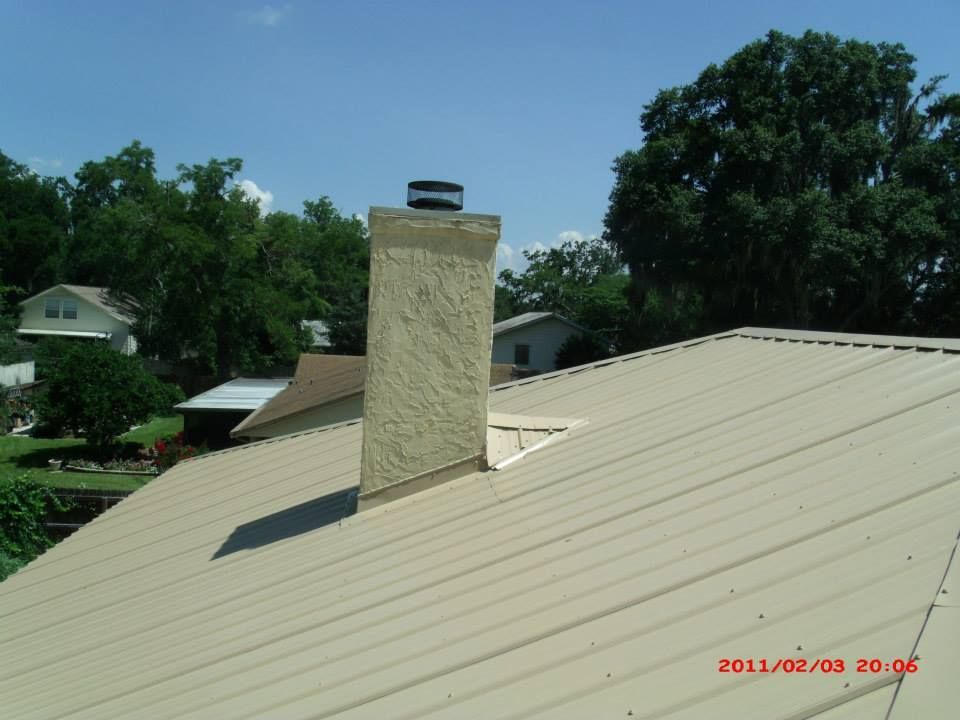 Chimney on a tan metal roof with a blue sky background and trees.