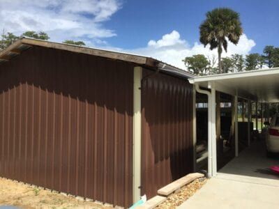 Brown metal-sided shed next to a carport. Blue sky with palm tree in the background.