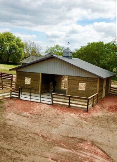 Wooden horse barn with metal roof, light brown siding, and fenced-in area under a cloudy sky.