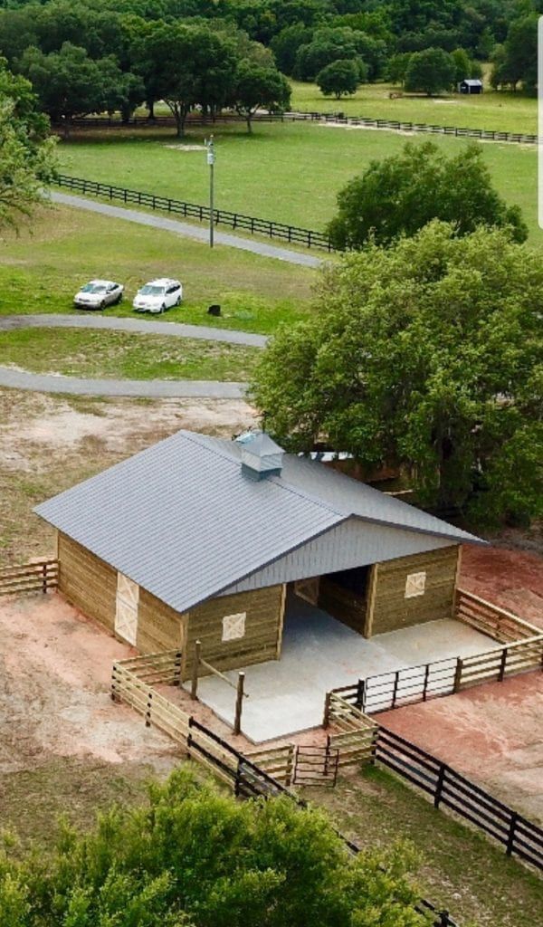 A rustic barn with a gray metal roof, surrounded by a fence and open fields. Two white cars parked nearby.