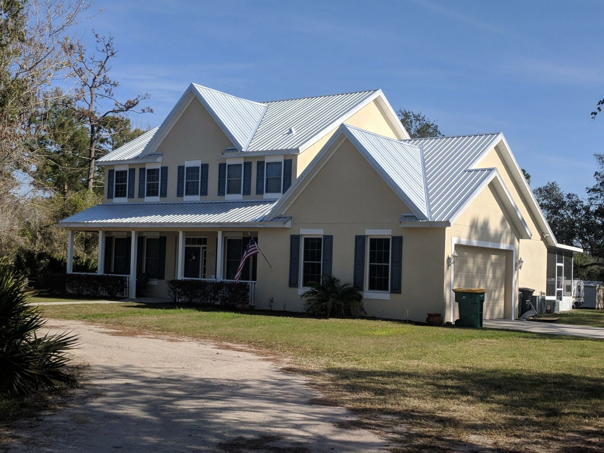 Two-story tan house with gray shutters and metal roof, porch, and attached garage on a grassy lot.