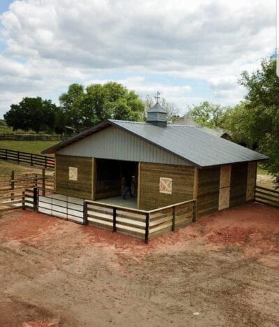 Wooden barn with metal roof, small porch, and surrounding fence, in a rural setting.