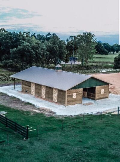 Horse barn with green roof, wooden walls, and concrete foundation, surrounded by green grass and trees.