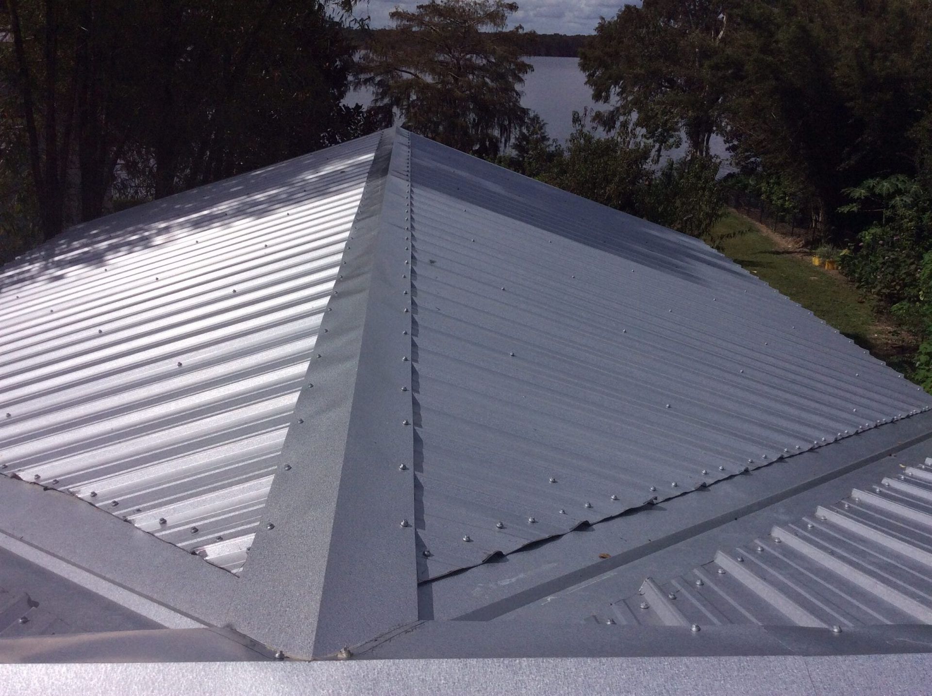 Gray metal roof with pyramid-shaped peak. Surrounded by trees and a body of water.