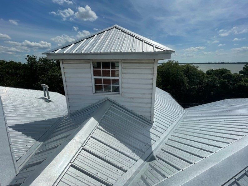 Metal roof with white cupola and window, blue sky, trees, and water in background.