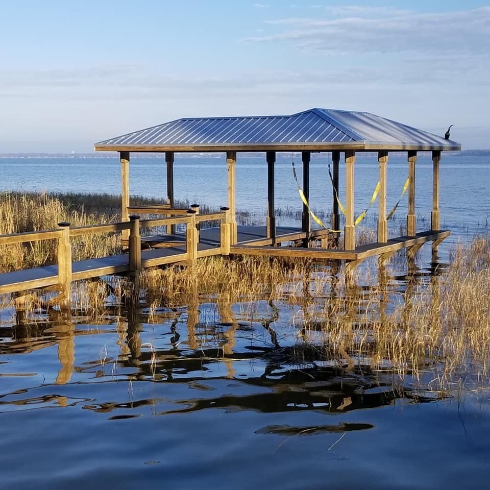 Wooden gazebo on a dock over water, reflected in the ripples. Tall grass frames the foreground, calm blue sea in the background.