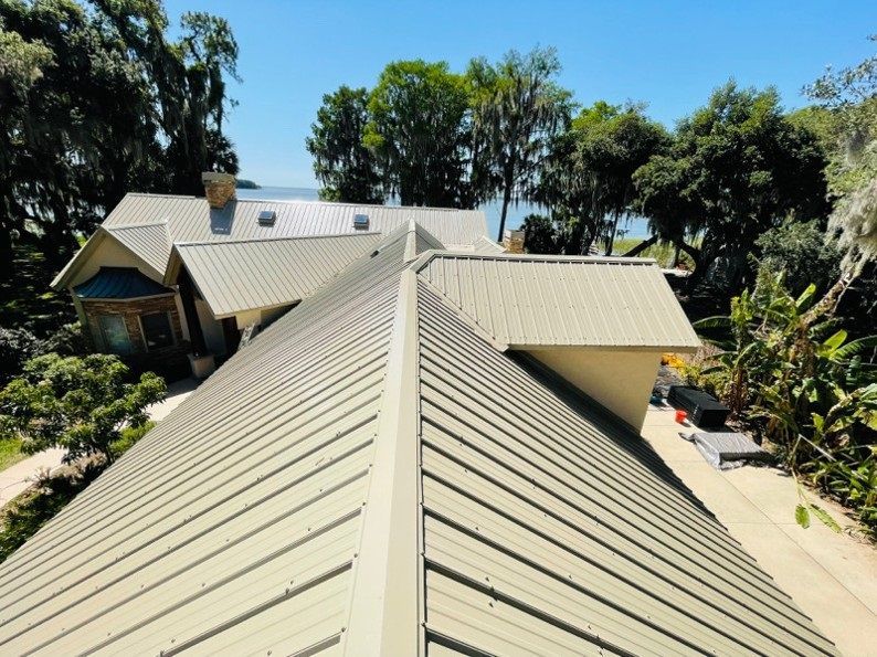 Aerial view of a house with a metallic green roof on a sunny day, trees and water in the background.