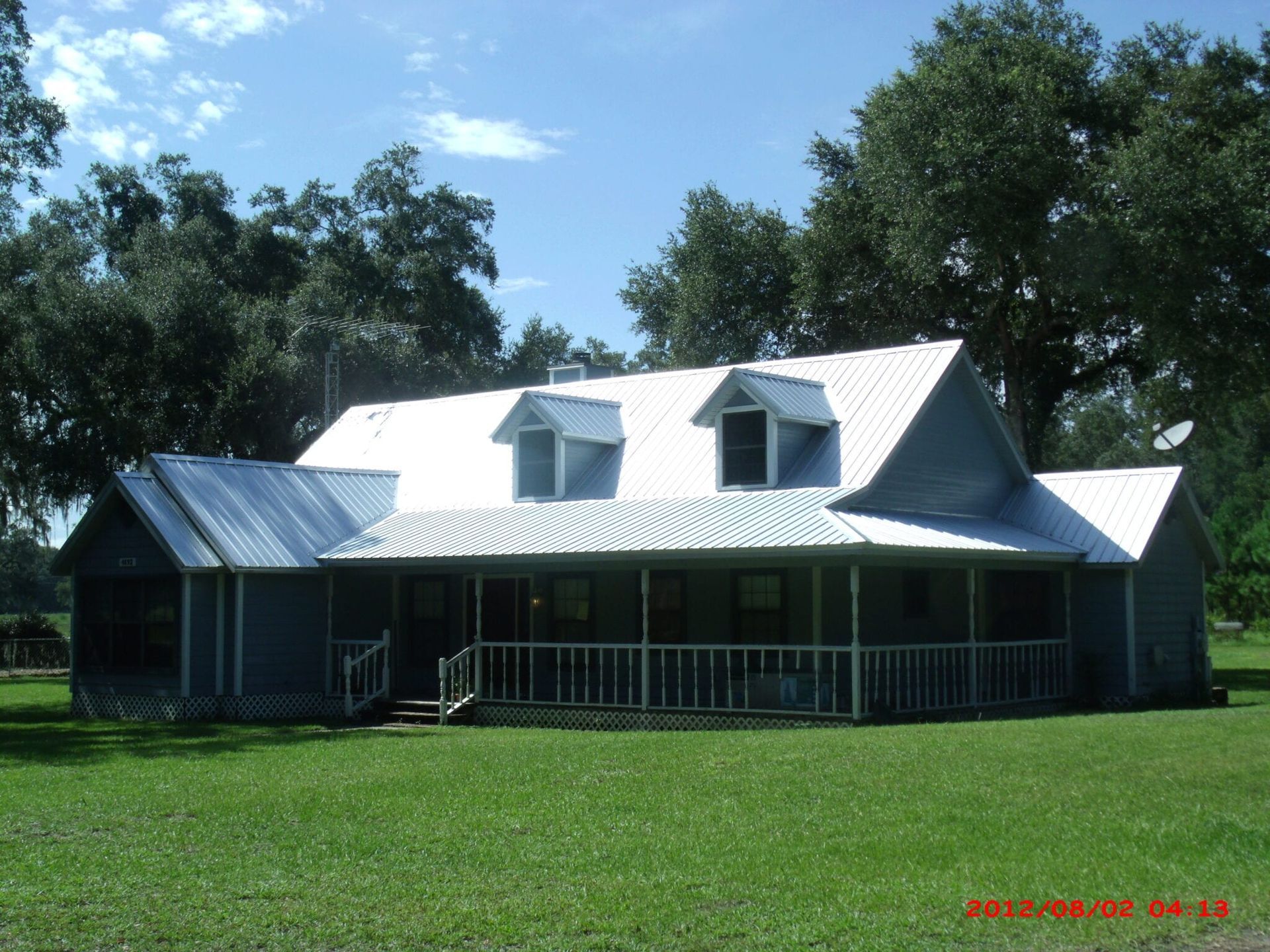 Blue house with white roof, porch, and dormers. Green lawn, trees, and blue sky in background.