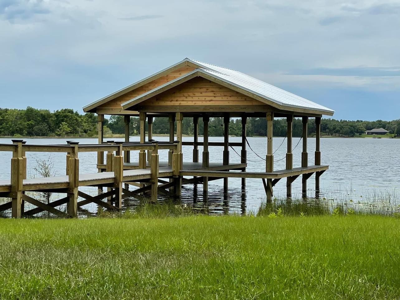 Wooden boathouse with a metal roof on a lake, green grass in the foreground.