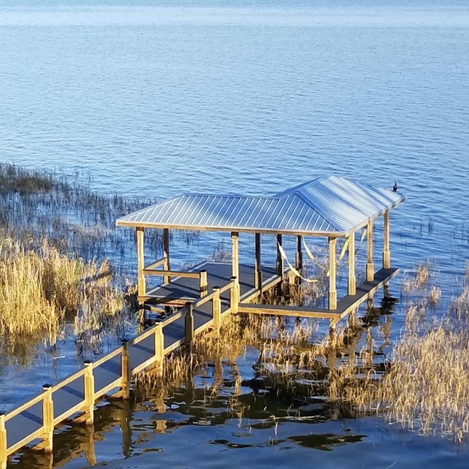 Wooden dock with a covered seating area on a lake with tall reeds. Blue water and sky.