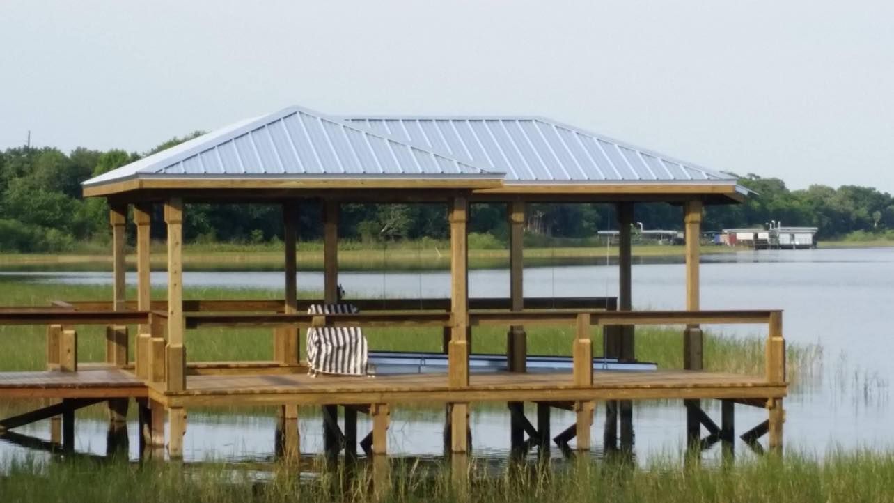 Wooden gazebo with metal roof on a pier over a lake, surrounded by marsh grass.