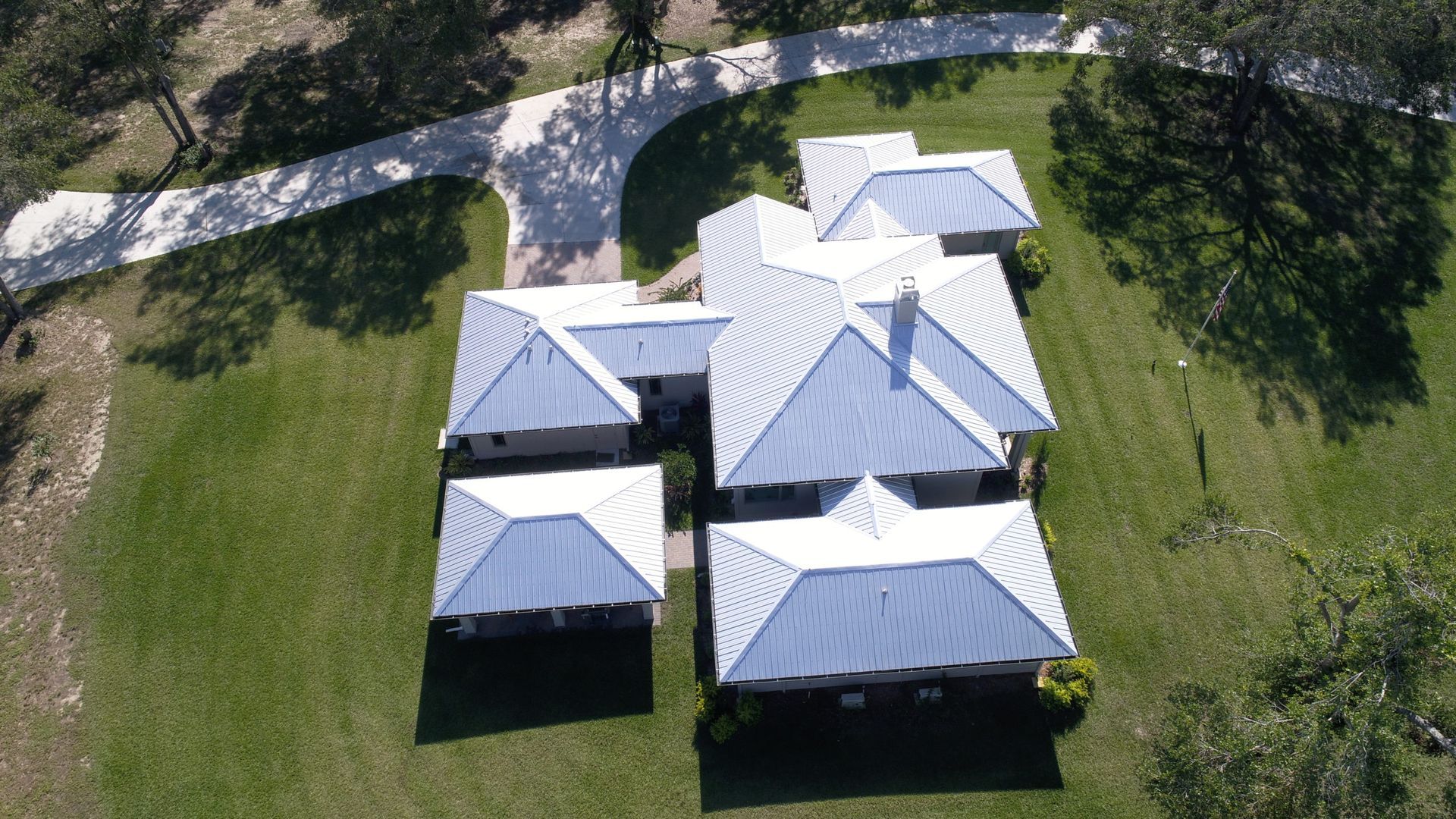 Aerial view of a white-roofed house with a winding driveway, surrounded by green grass and trees.