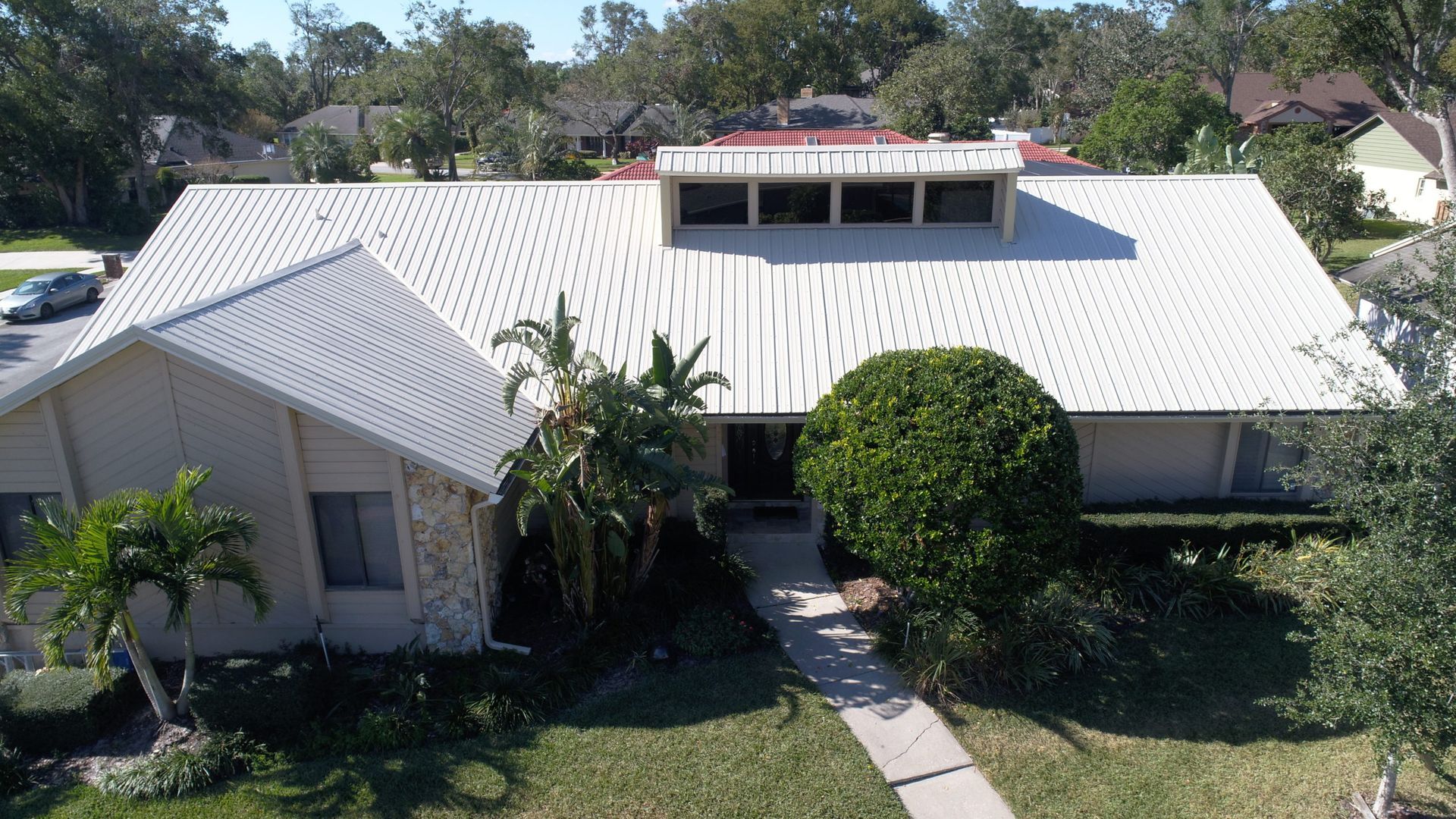 House with white metal roof and light-colored exterior, surrounded by green grass and trees.