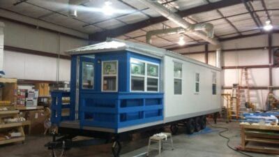 A blue and white tiny home on wheels inside a workshop, with a porch and multiple windows.