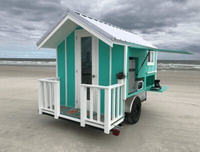 Teal tiny house on wheels with white trim and porch on a beach.