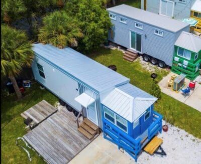 Aerial view of a blue and gray tiny home with a deck, palm trees, and other houses.