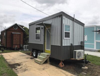 Tiny house with metal siding, gray and white, parked on a concrete pad, next to a small wooden structure.