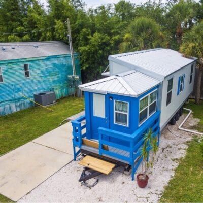 Tiny house with blue porch, white siding, parked next to a blue building, on gravel, with greenery.