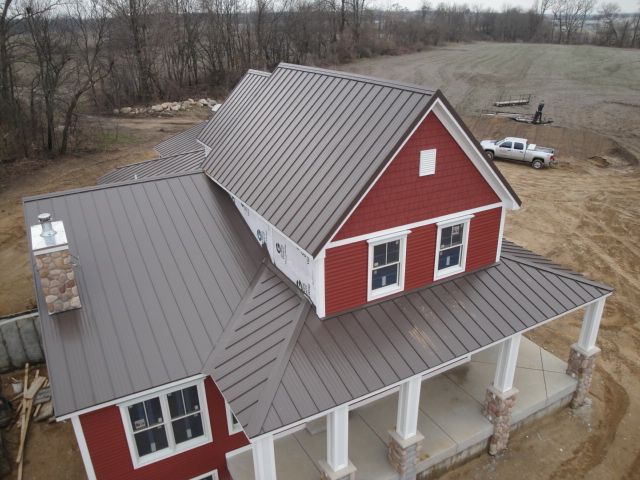 Red house with brown metal roof, porch, and white trim in rural setting.