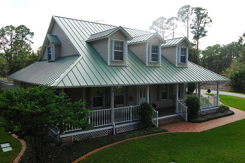 A light-colored house with a green metal roof and a wraparound porch, surrounded by green grass and trees.