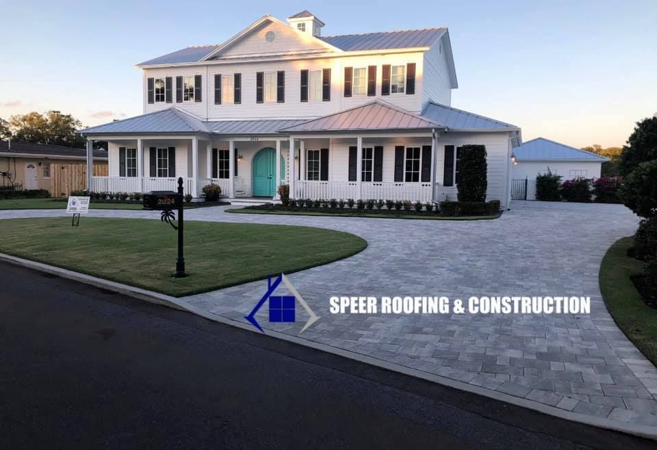 White house with blue door, gray paver driveway, and green lawn; advertisement for Speer Roofing & Construction.